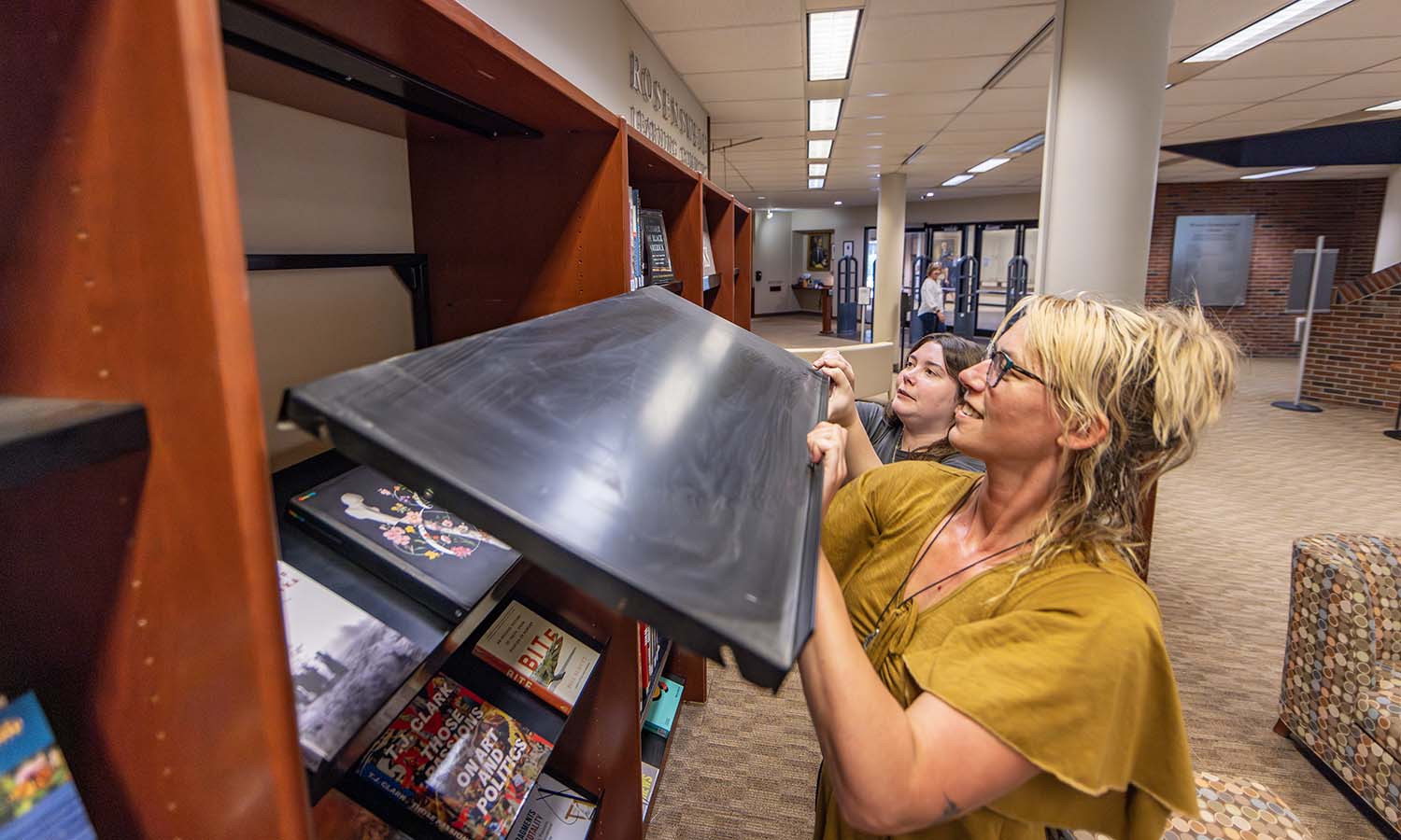 In the Rosensweig Learning Commons of the Warren Hunting Smith Library, Access Services Specialist Catherine Morse-Jahna and Access Service Coordinator Lisa Gibson clean and organize shelves to display books by faculty authors. 