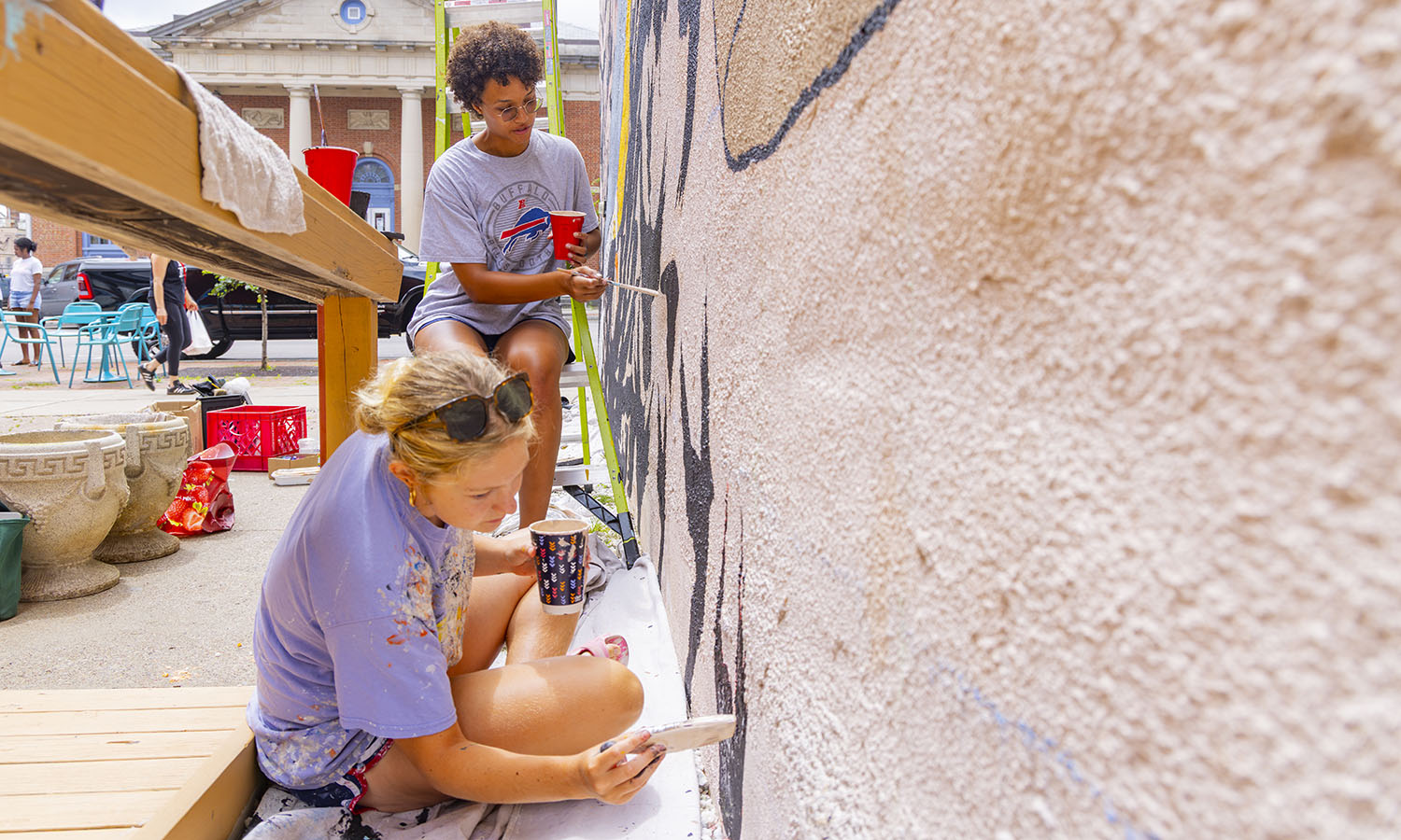 Ella Rhodes '29 and Nora Smith '27 paint a mural, designed by artist Tsioianiio Galban, on Castle Street in downtown Geneva. Rhodes and Smith are working with Associate Professor of Gender, Sexuality, and Intersectional Justice Michelle Martin-Baron on PACT, the Public Art for Community Transformation Summer Mural Project. 